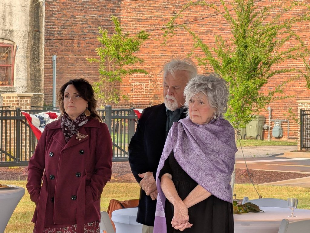 Three people stand outdoors near tables, looking serious. Two older adults and one younger woman are dressed warmly. There is a brick wall and tree in the background.