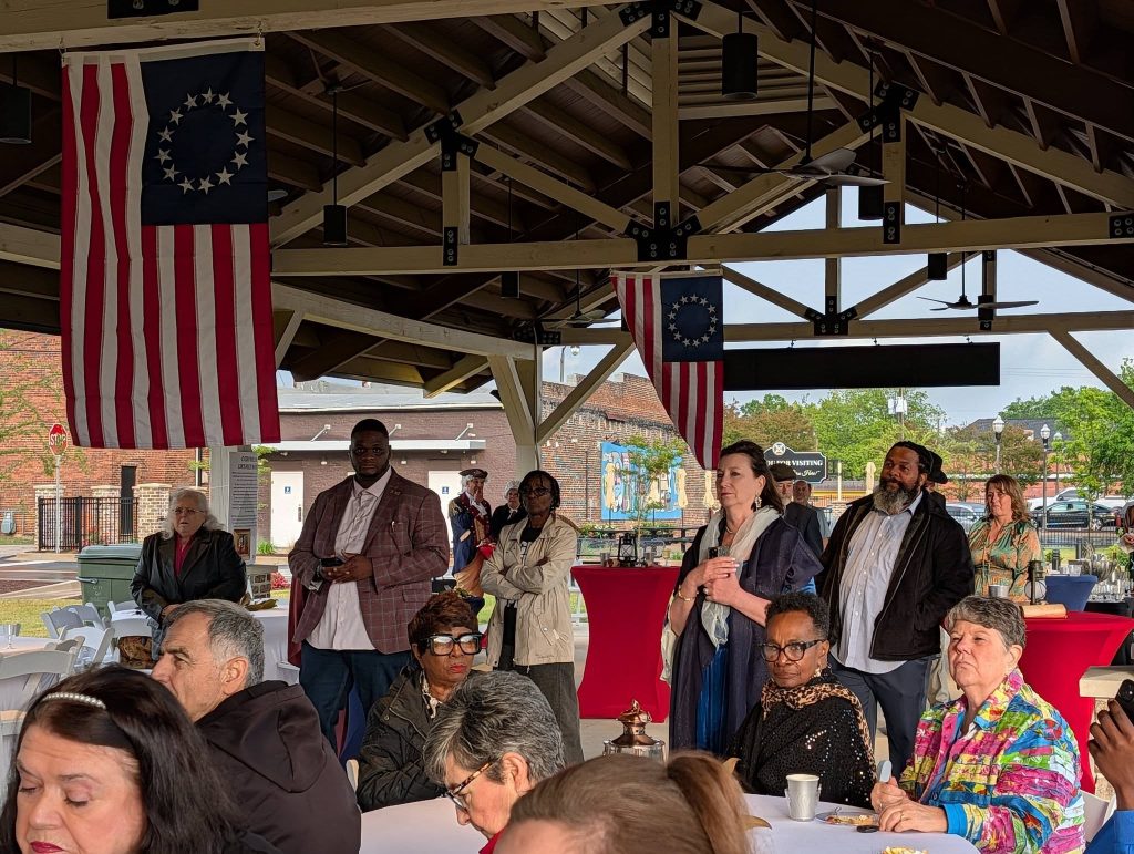 People gathered under a pavilion with colonial American flags, some sitting at tables and others standing, engaged in what appears to be a formal or commemorative event.