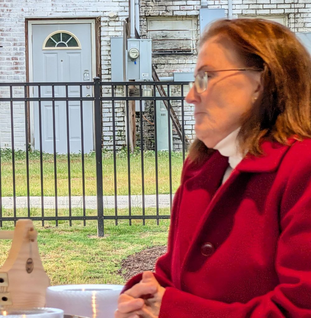 Woman in a red coat sits at a table outdoors, with a blurred background of a white brick building, gate, and grassy area.