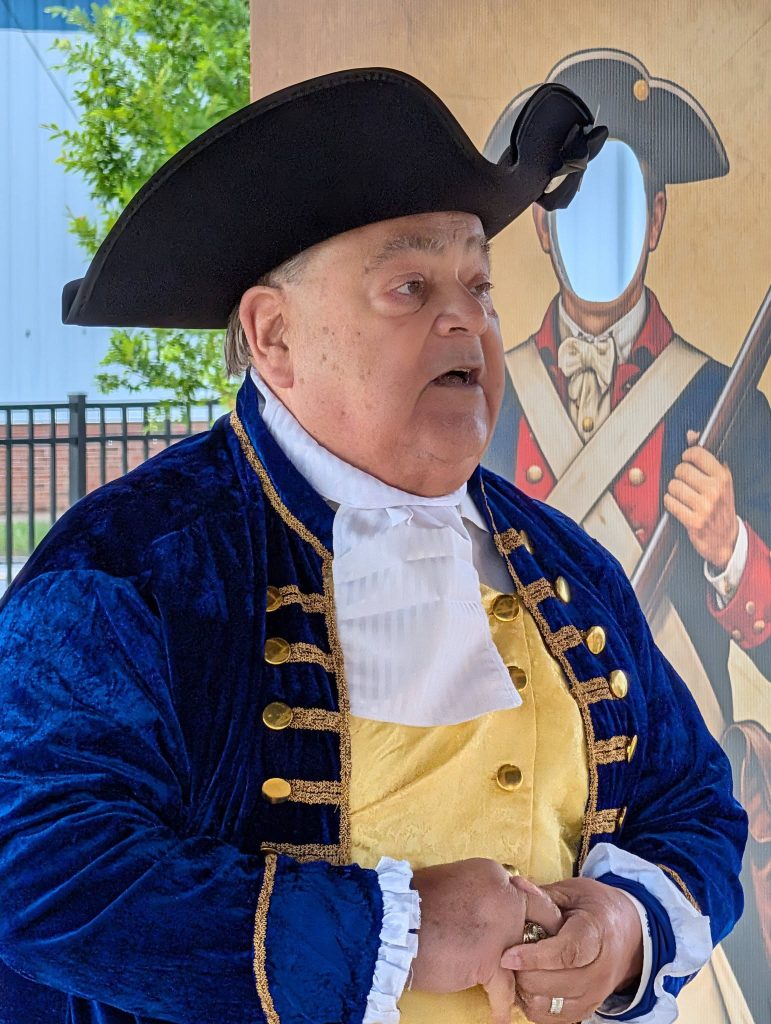 older man in a colonial costume with a tricorn hat stands in front of a faceless soldier cutout and a fence, outdoors.