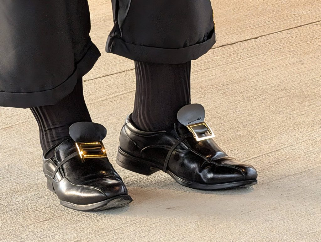 Person wearing shiny black shoes with large gold square buckles, black socks, and black pants with rolled cuffs, standing on a textured concrete surface.
