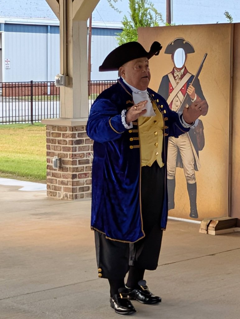 Man in colonial-era costume speaks outdoors, standing in front of a cutout soldier display, under a shelter with brick and wooden supports.