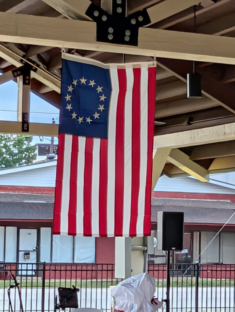 The Betsy Ross American flag with thirteen stars in a circle hangs under a pavilion roof.