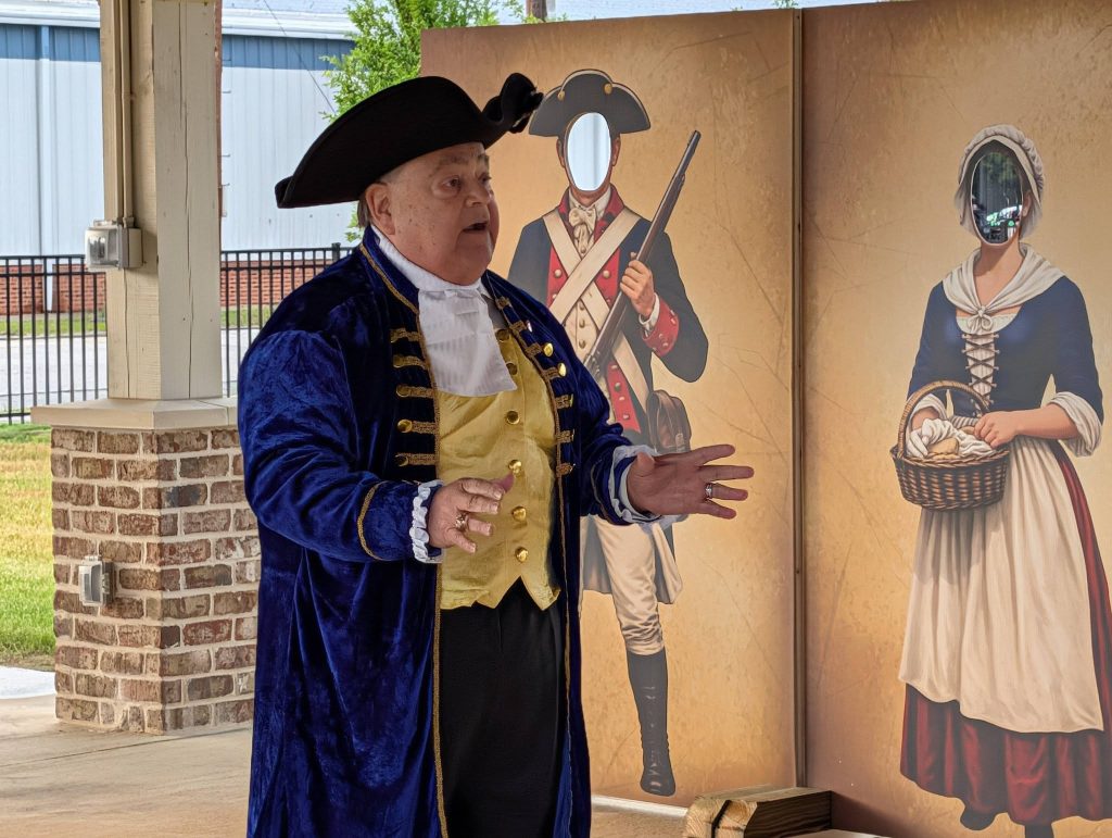 older man in colonial attire speaks indoors, with historical costume boards for people to pose behind in the background.