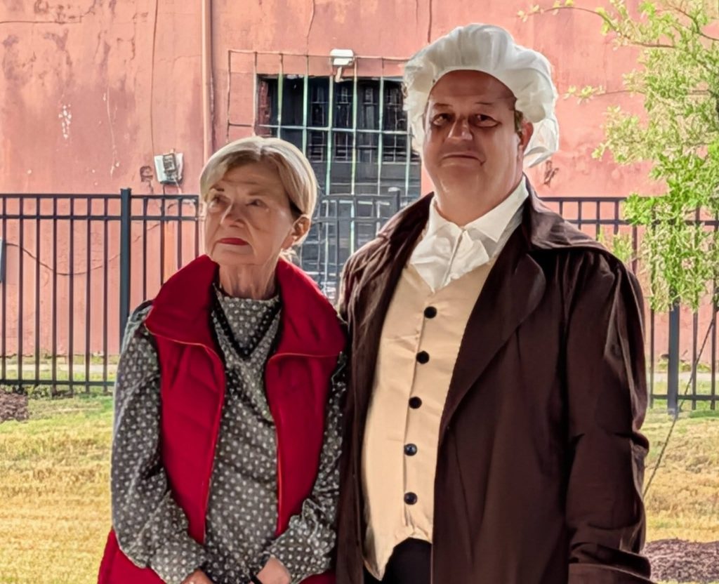 Two adults pose outdoors, one in a red vest and polka dot shirt, the other in a colonial-era costume with a white wig and brown coat.