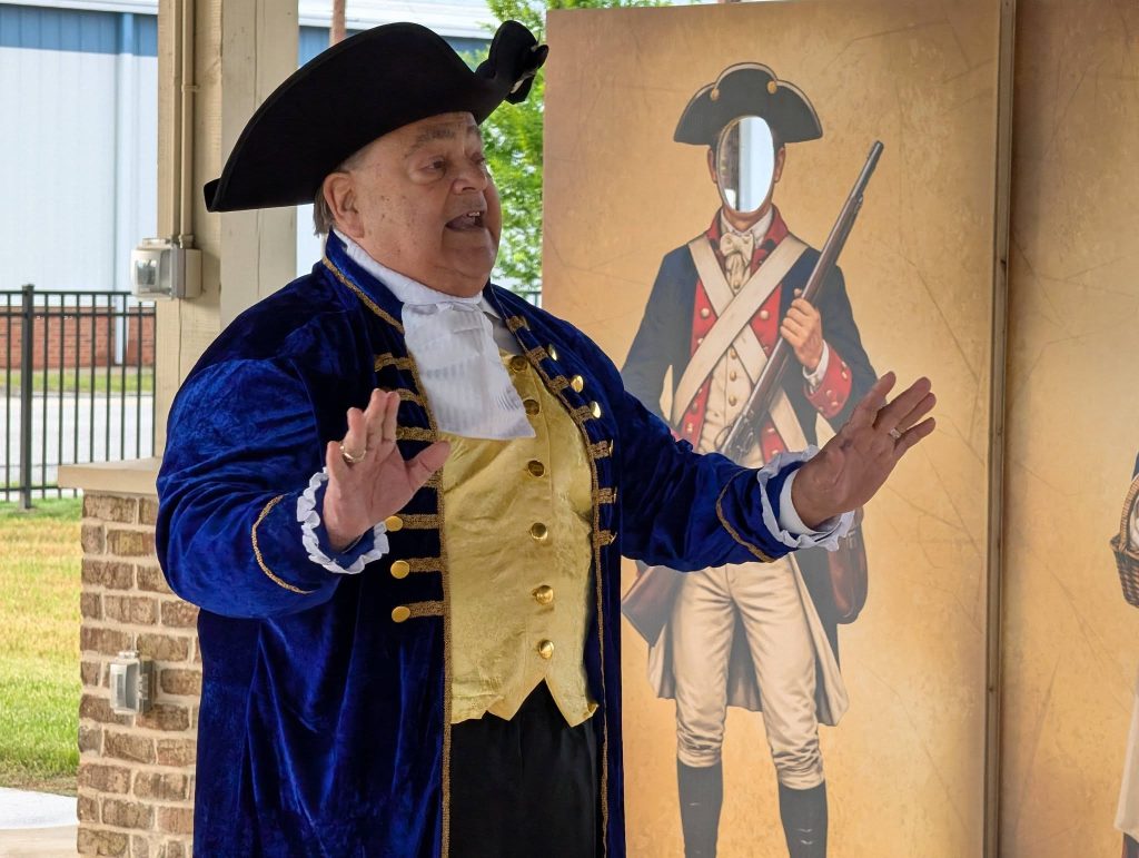 older man in colonial attire speaks in front of a standee depicting a historical soldier with a cutout for a face.