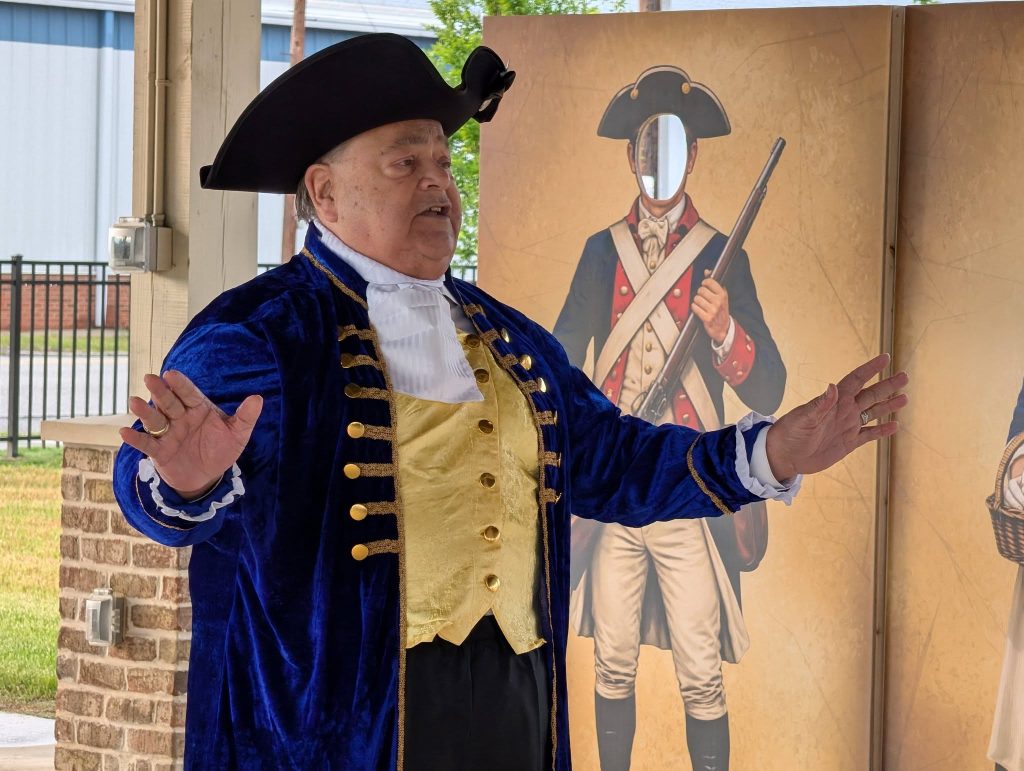 Man dressed in colonial-era costume passionately speaking, with hands raised. Behind him is a standee of a historical soldier with a face cutout for photos.