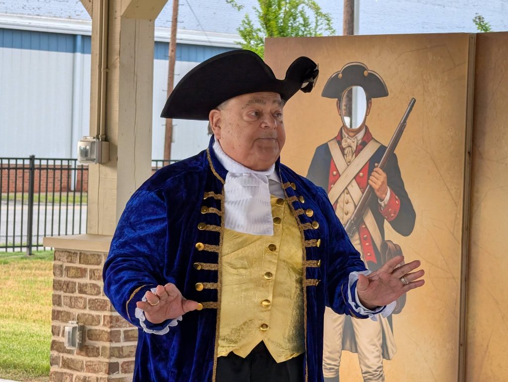 Older man in colonial costume stands indoors, gesturing, with a cutout of a revolutionary soldier with a face hole in the background.