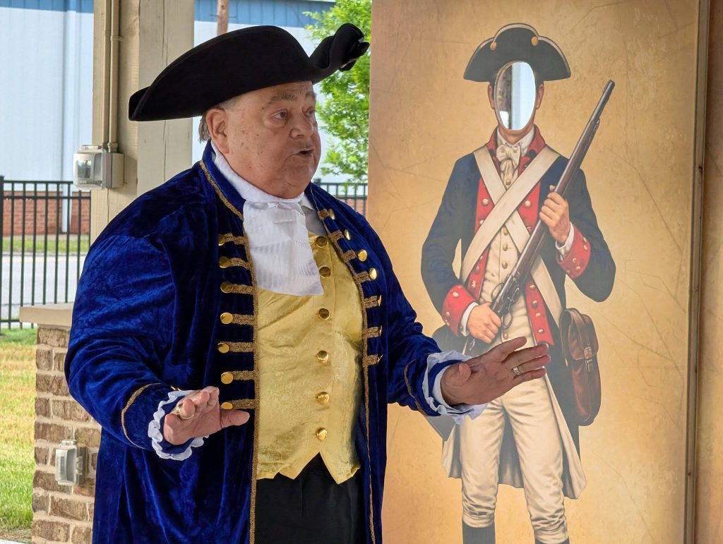 Man in colonial costume gestures while standing next to a standee of a historical soldier with a cutout for the face.