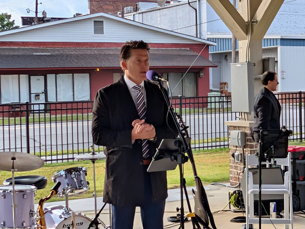 A man in a suit speaks into a microphone at an outdoor event. Musical instruments and another person are visible in the background.