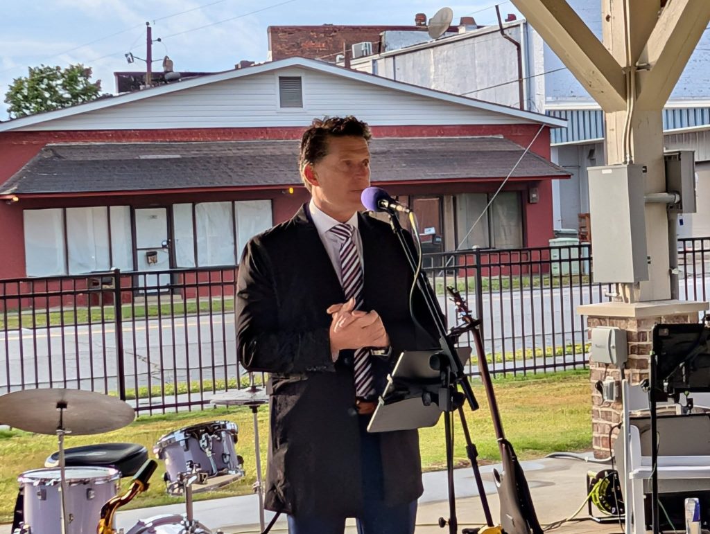 A man in a suit speaks into a microphone at an outdoor event, with musical instruments and buildings in the background.