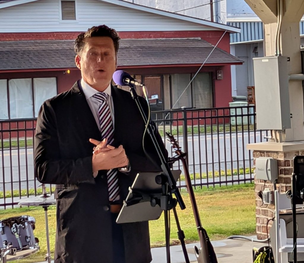 Man in a suit speaking into a microphone outdoors, standing near musical instruments and equipment, with buildings and a fence in the background.