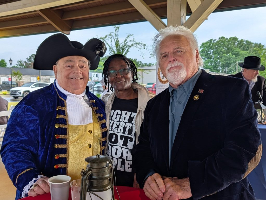 Three people pose together at an outdoor event, one wearing colonial attire, another in casual clothing, and the third in a suit jacket with pins.