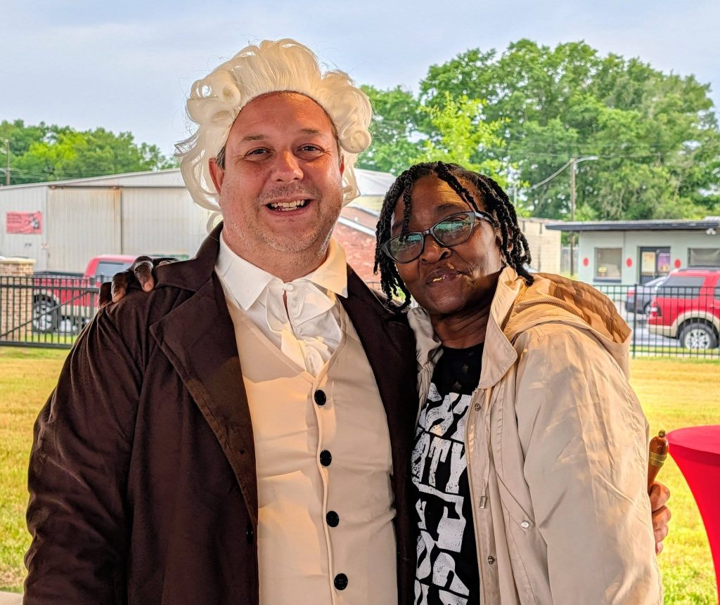 Two people standing together outdoors, one wearing a historical costume with a white wig, and the other in casual clothing and glasses, smiling at the camera.