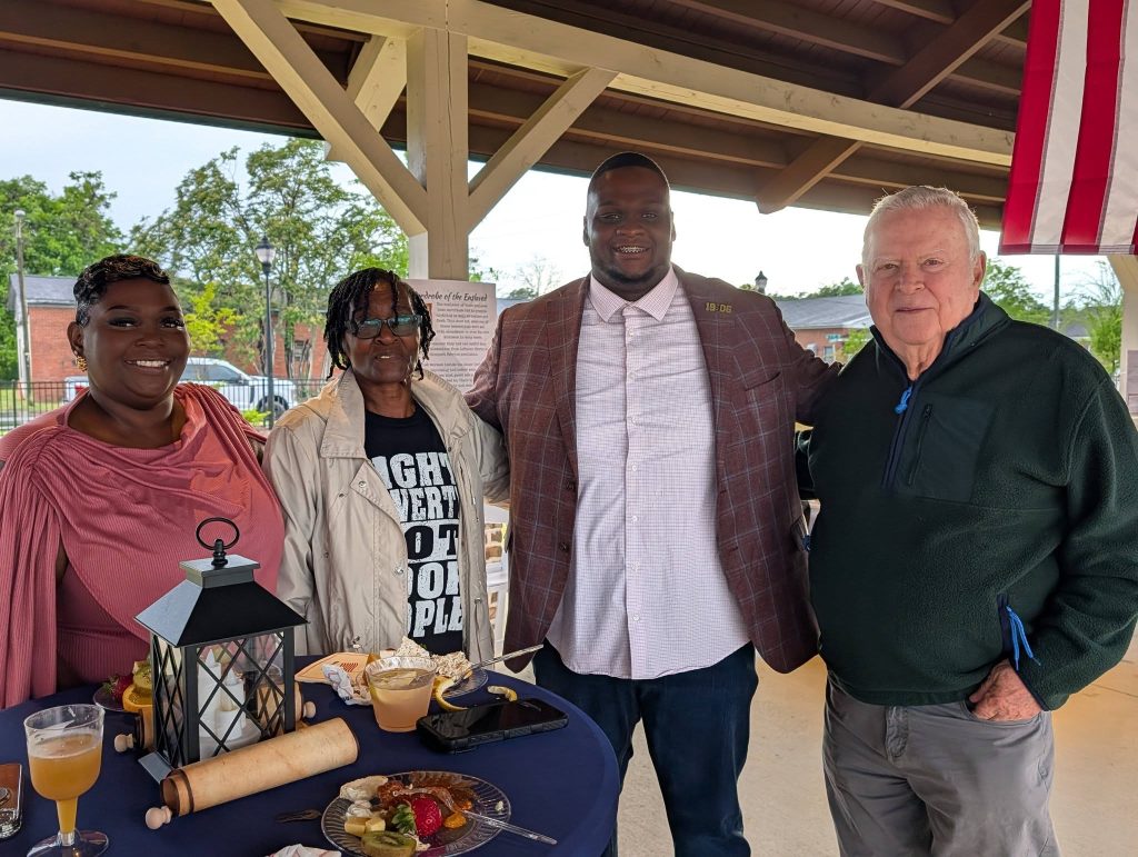 Four people smiling and posing together at an outdoor covered event, standing behind a table with food, drinks, and a lantern centerpiece.
