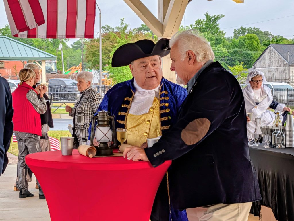 Two men, one dressed in a historical costume, converse at a red table under a pavilion, with others in colonial attire and modern clothing in the background.