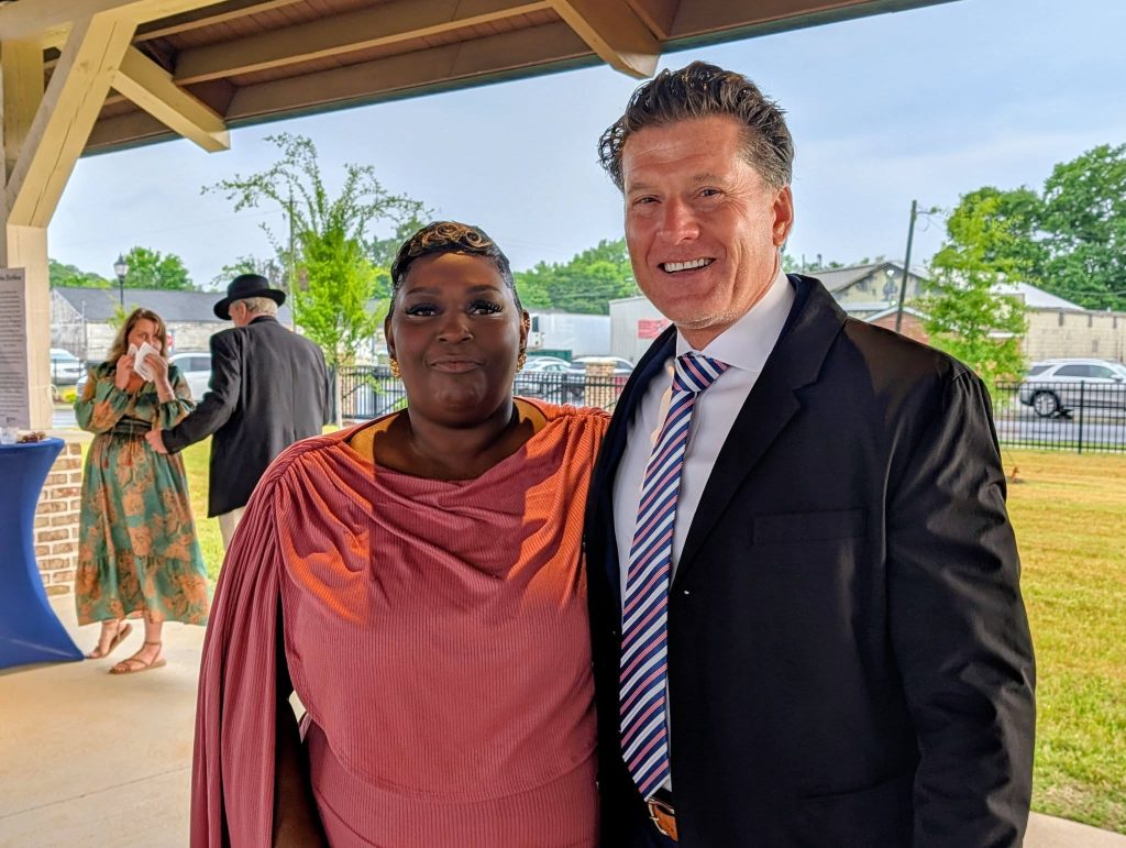 A woman in a pink dress and a man in a suit smile together at an outdoor event, with two more people in the background.