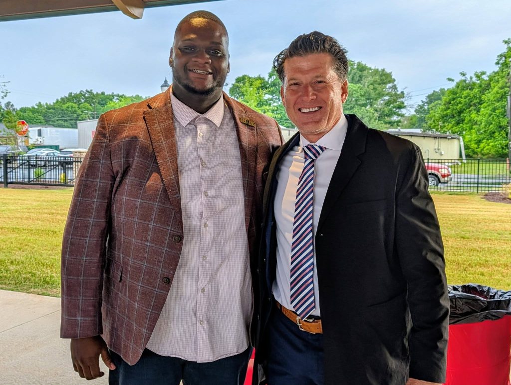 Two men dressed in suits stand side by side outdoors, smiling at the camera with trees, grass, and a street visible in the background.