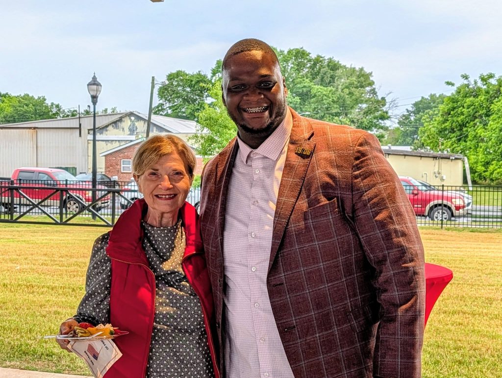 older woman and a younger man smiling together outdoors. She holds a plate of food, both are dressed nicely, and cars and trees are visible in the background.