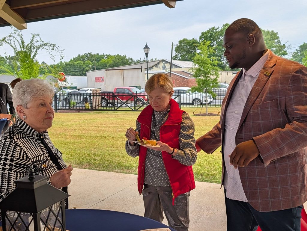 Three adults interact outdoors under a pavilion; one woman holds a plate of food, while the others engage in conversation on a sunny day.