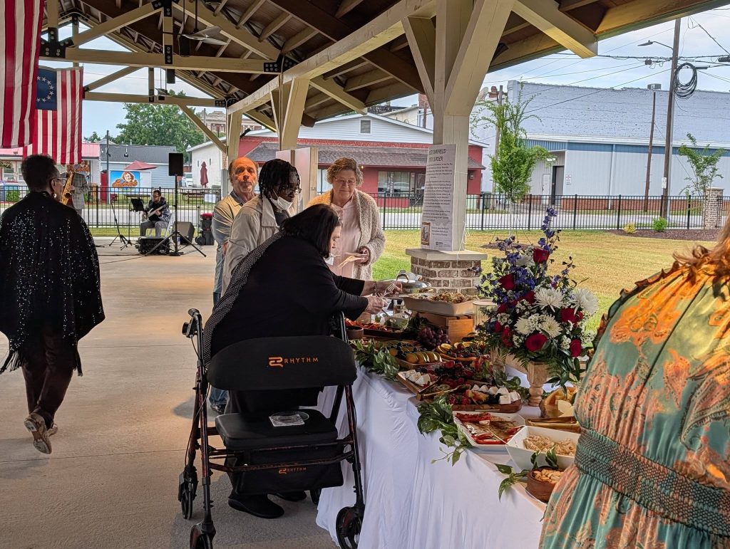 People gathering under a pavilion, serving food from a decorated buffet table with flowers, while a musician plays guitar in the background, and an American flag hangs overhead.