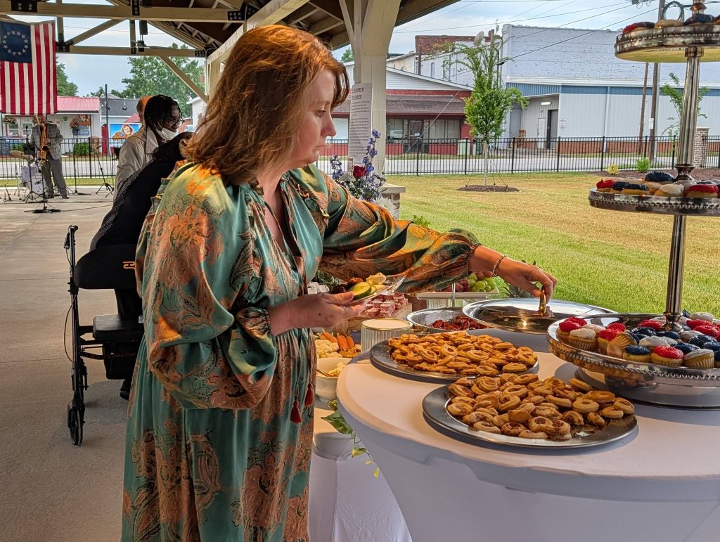 A woman in a patterned dress selects pastries from a buffet table at an outdoor event, with musicians and an American flag visible in the background.
