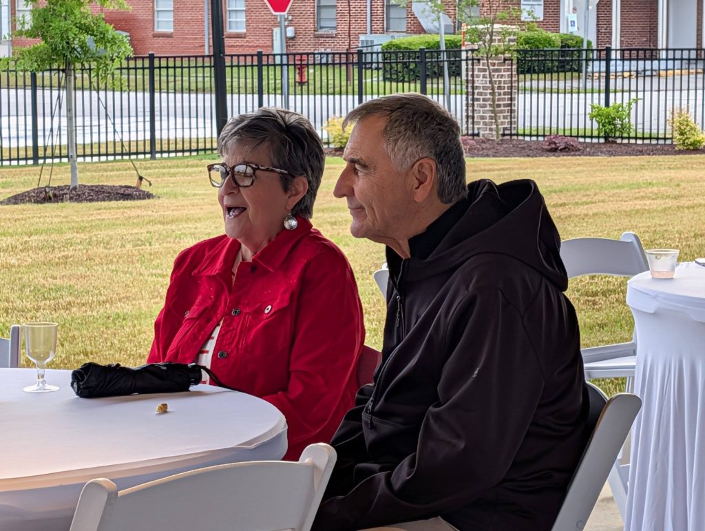 older woman and man sit together at a round table outdoors, smiling and enjoying an event under a tent.