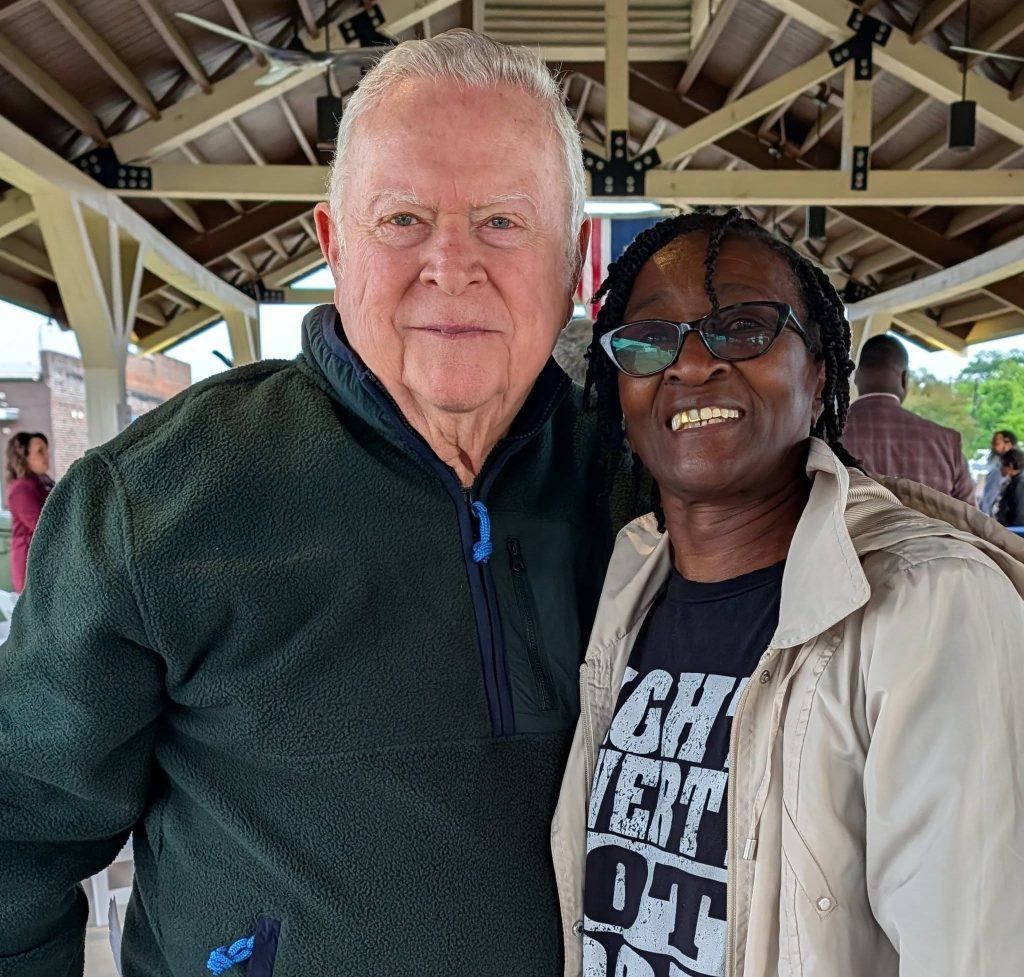 older man and woman smiling together under a pavilion, with people and outdoor scenery in the background.