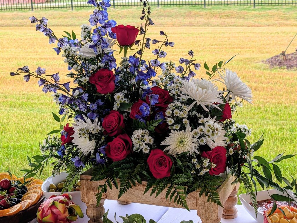 A wooden box filled with red roses, white chrysanthemums, purple flowers, and green leaves, displayed on a table with fruit in the background.