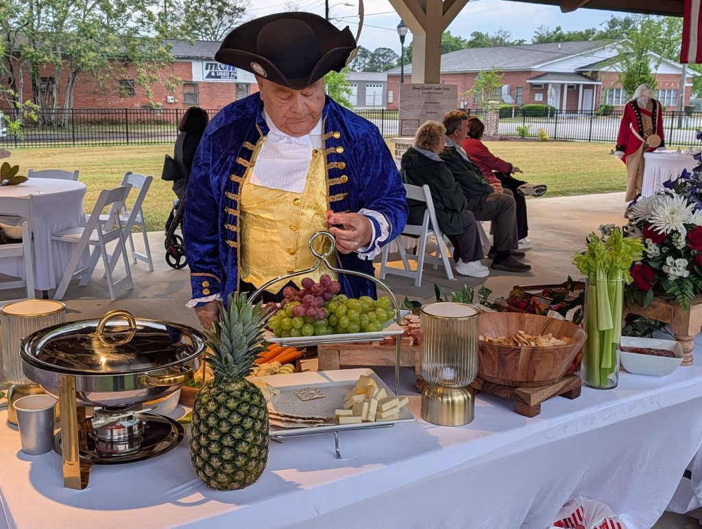 Man in historical costume stands at a table with fruit, cheese, pineapple, and snacks during an outdoor event with other people seated in the background.