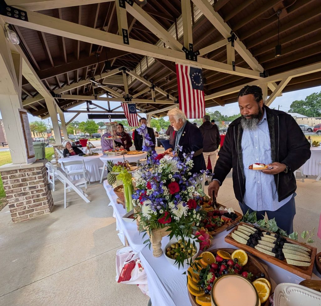 People gather under a pavilion decorated with American flags, enjoying a buffet table filled with flowers, fruits, and desserts during a social event.