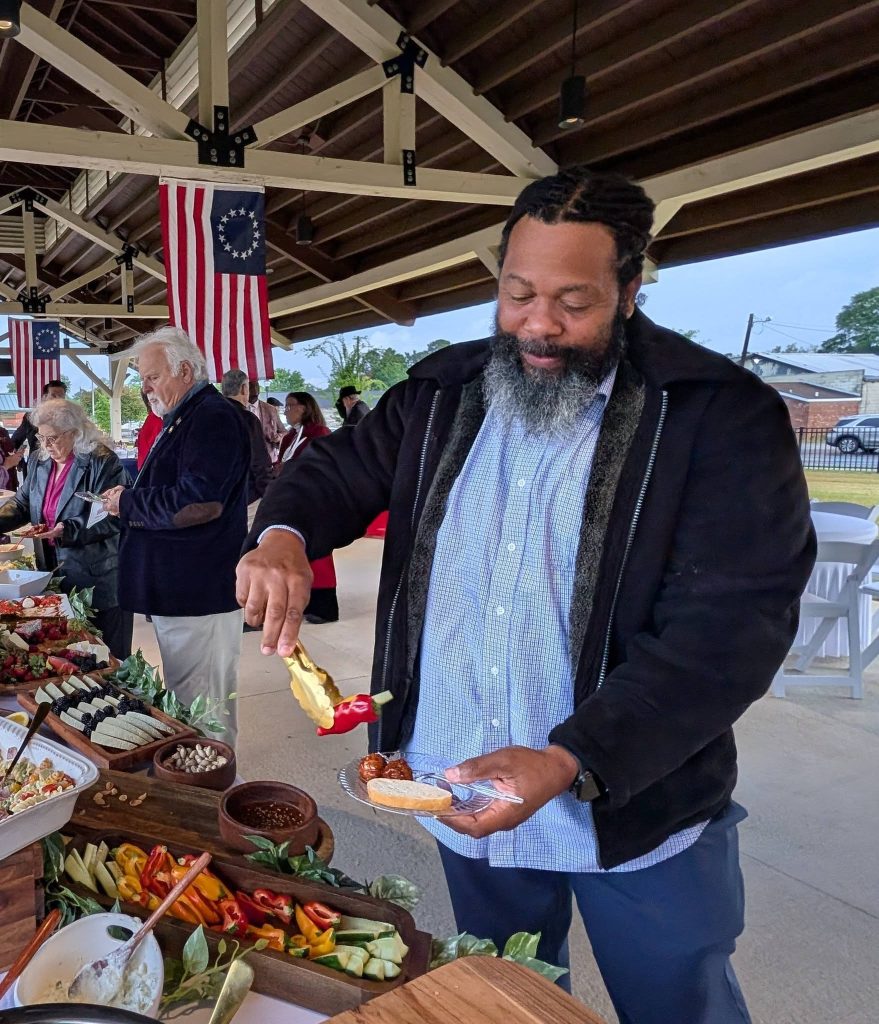 A man serves himself food at a buffet table, under a pavilion decorated with American flags, while other people are also getting food in the background.