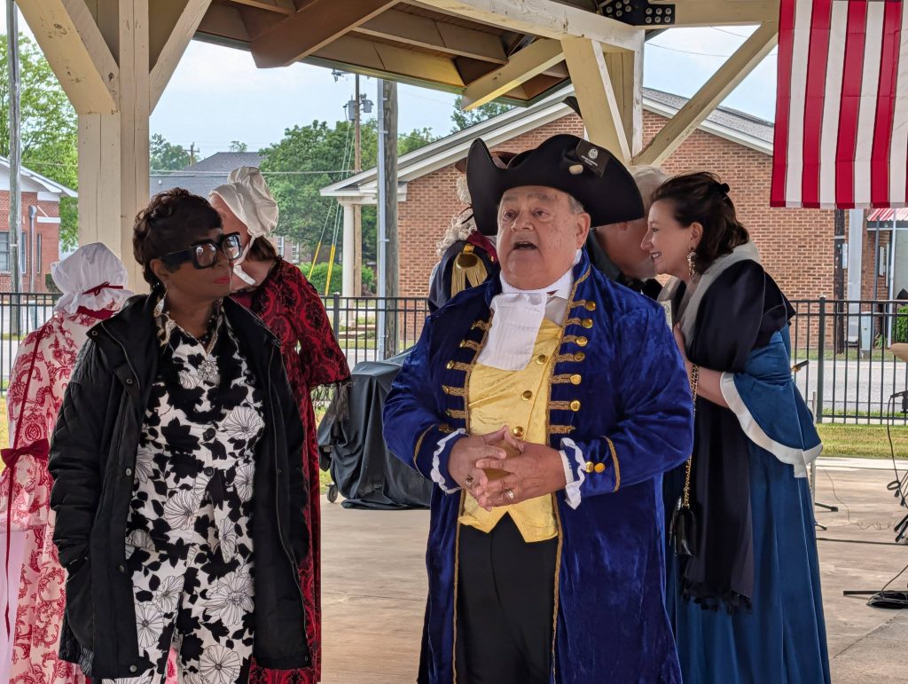 Several people dressed in colonial-era costumes stand under a pavilion, with one man in a blue and yellow outfit speaking, and an American flag hanging in the background.