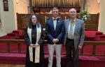 Three people pose inside a church; a woman in clergy attire, a young man in a suit, and an older man in a suit, with organ pipes in the background.