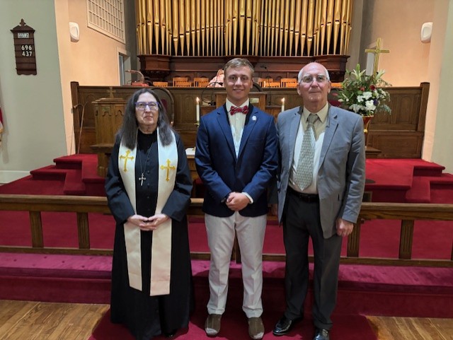 Three people pose inside a church; a woman in clergy attire, a young man in a suit, and an older man in a suit, with organ pipes in the background.
