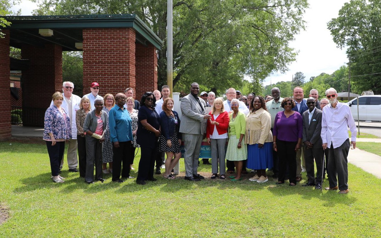 A diverse group of adults poses together outdoors on grass in front of a brick structure, smiling at the camera under a sunny sky with trees in the background.