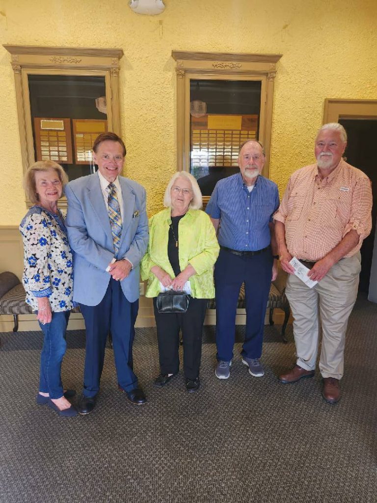 Five older adults standing together indoors, smiling at the camera, with a yellow wall and framed windows in the background.