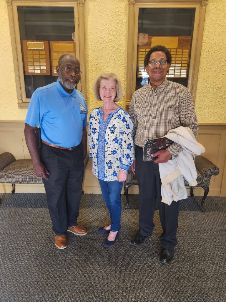 Three adults stand smiling indoors between two benches, posing for a photo. The setting has yellow walls and framed plaques in the background.