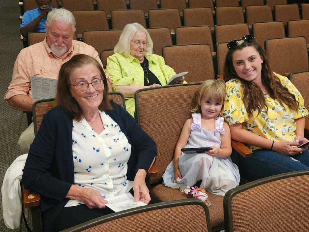 Five people of different ages sitting in an auditorium, smiling and holding papers or devices, with many empty brown seats around them.