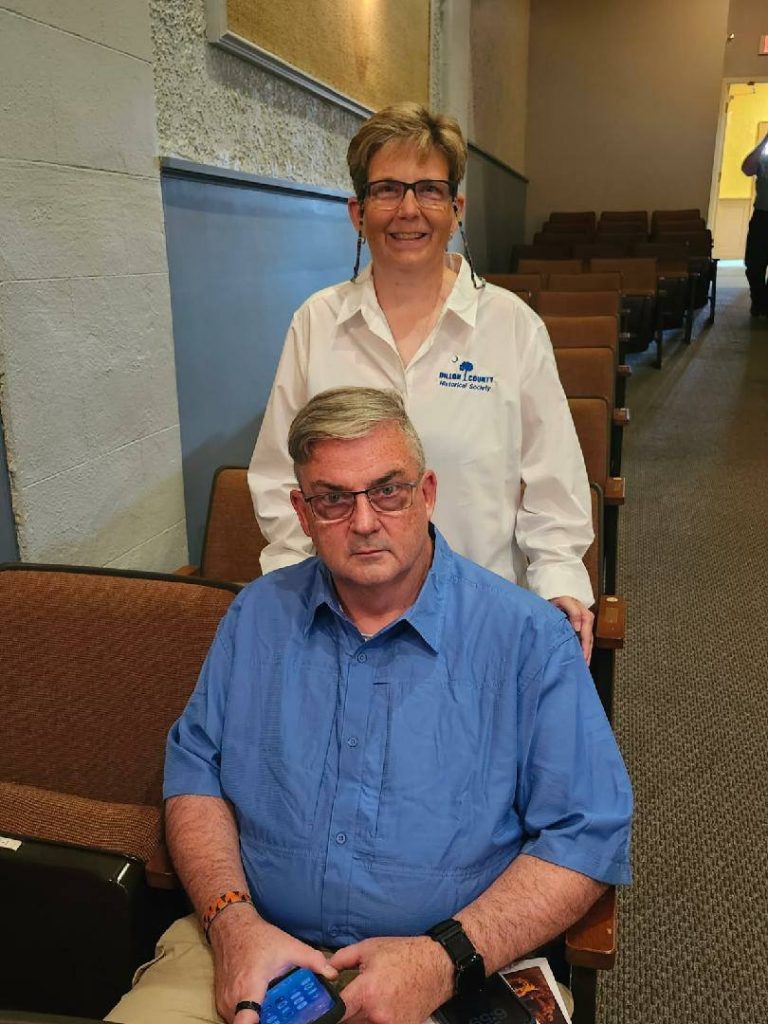 Two adults in a theater or lecture hall, one seated in front holding a phone, and one standing behind, both wearing glasses and casual clothes.