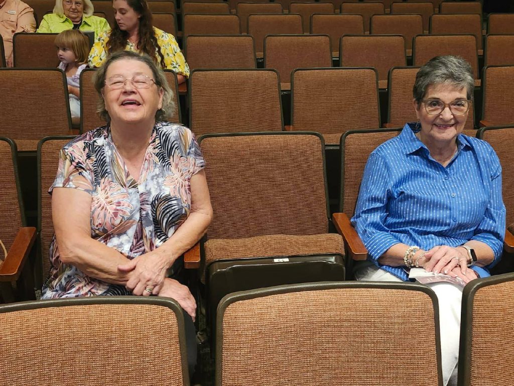 Two elderly women sitting and smiling in a mostly empty auditorium with brown seats, while a few other people sit in the background.