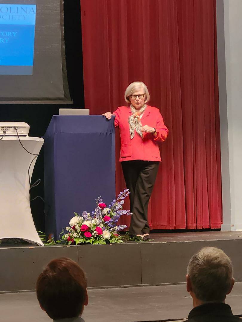 A woman in a red jacket stands on stage beside a podium and floral arrangement, speaking to an audience with a red curtain backdrop.