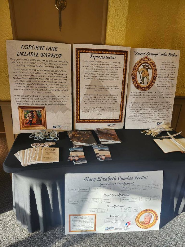 A display table with historical information boards, booklets, pamphlets, wristbands, and a genealogy chart about Mary Elizabeth Cumbee Freitas and family history.
