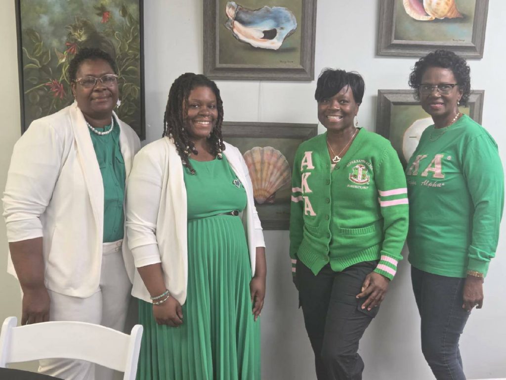 Four women wearing green and white outfits pose together, smiling, in front of framed shell and flower artwork on a wall.