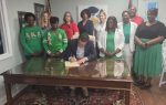 A man sits at a desk signing papers while a group of people, some wearing green AKA attire, stand behind him, posing for a photo.