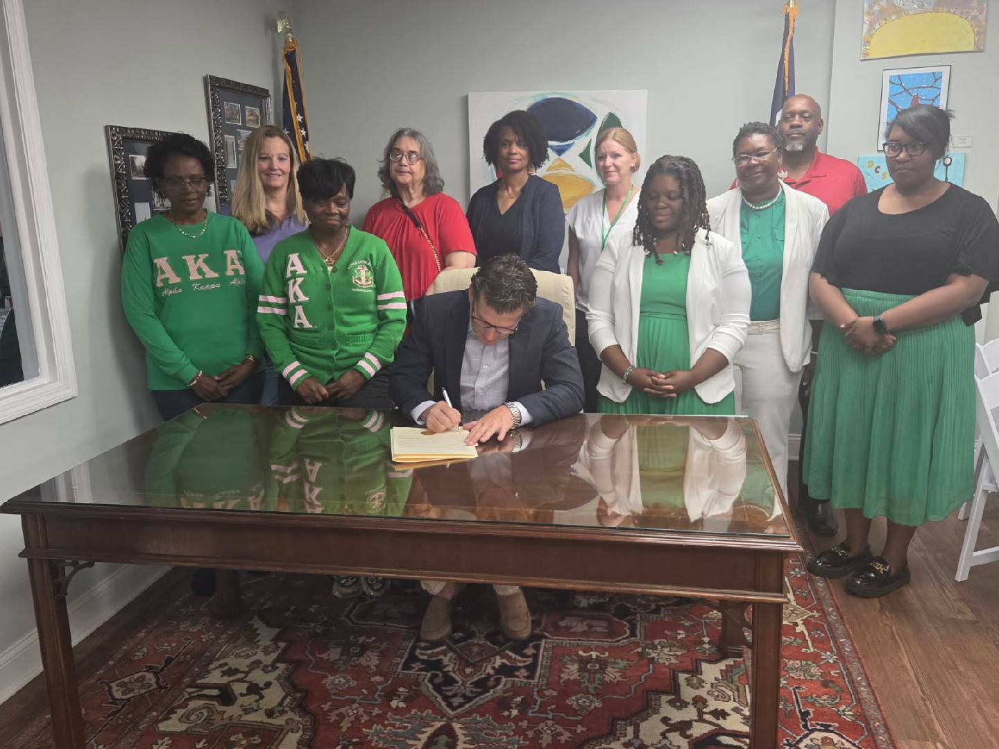 A man sits at a desk signing papers while a group of people, some wearing green AKA attire, stand behind him, posing for a photo.