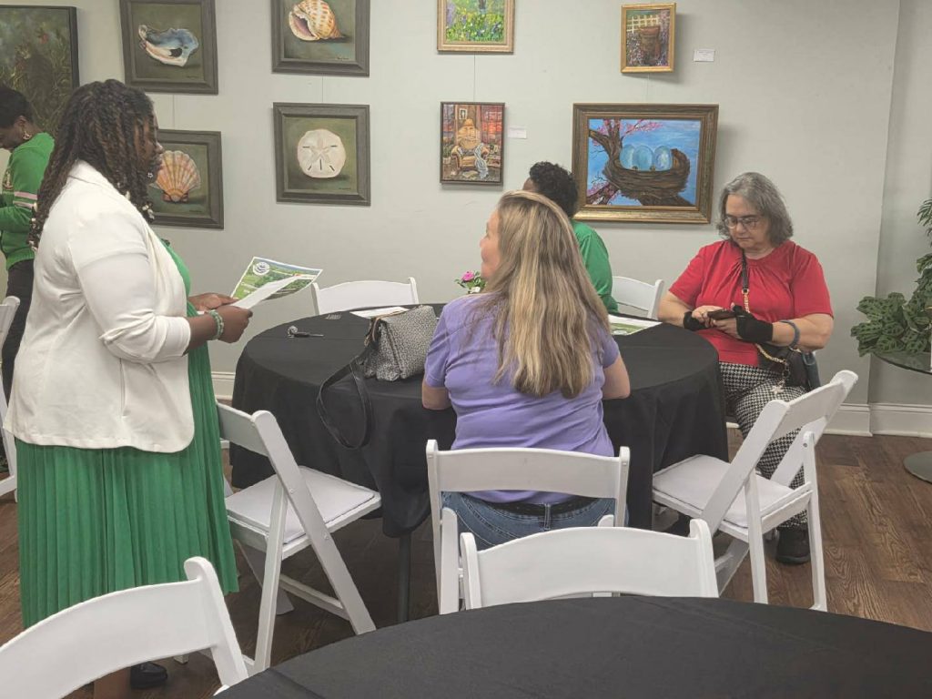 Four women sit and stand around a black-covered table, conversing and reading papers, with colorful artwork displayed on the walls behind them.