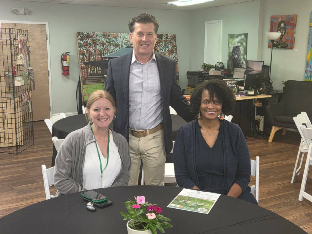 Two women seated at a round table with a man standing behind them in an office decorated with artwork and flowers.