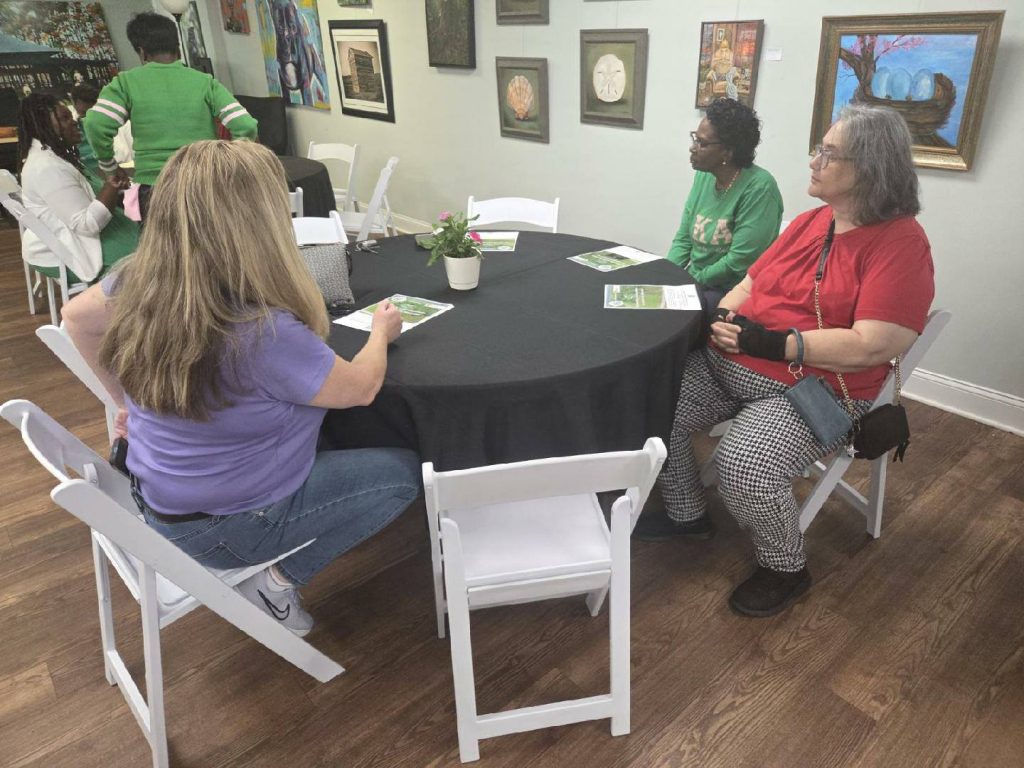 Three women sit at a round table in an art gallery, with paintings on the wall and informational papers on the table.