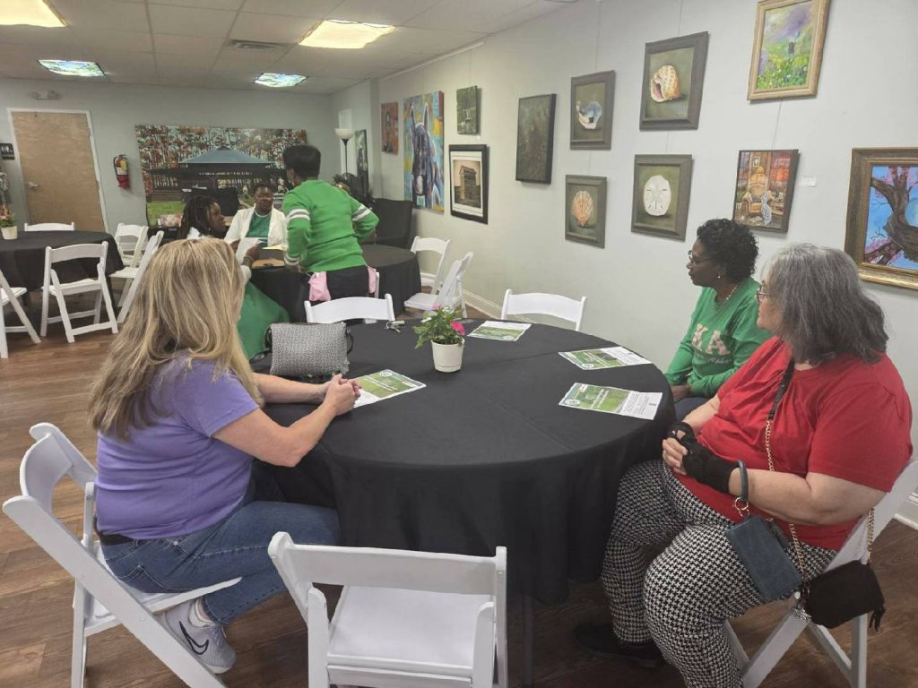 People sitting at round tables in an art gallery, chatting and reading papers, with paintings displayed on the wall in the background.